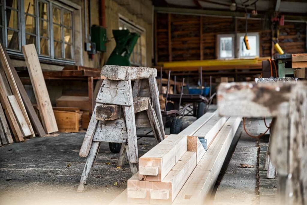 Eine Werkstatt mit Holzbalken und -brettern, einem verwitterten Sägebock, Werkzeugen und verstreutem Material. Der rustikale Raum mit abgenutzten Wänden und großen Fenstern spiegelt die Handwerkskunst von Baugeschäft und Zimmerei Müller Colditz wider.