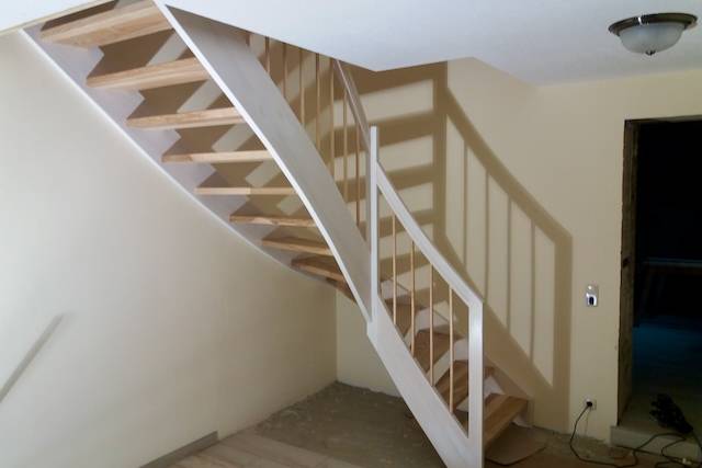 Eine moderne Holztreppe mit weißem Geländer schlängelt sich in einem gut beleuchteten Raum mit beigen Wänden und einer Deckenleuchte nach oben. Der Boden scheint unbearbeitet zu sein, und eine offene Tür führt in einen dunkleren Raum.