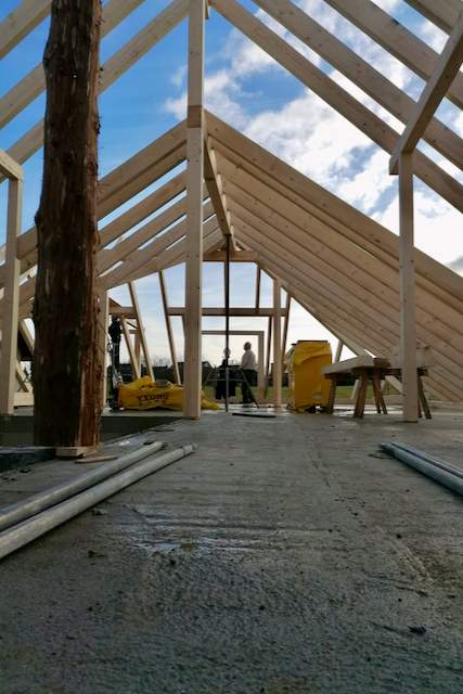 Ein teilweise gebautes Holzhaus mit freiliegenden Balken und einem wolkenverhangenen blauen Himmel darüber; auf dem Betonboden sind Baumaterialien und Werkzeuge zu sehen.
