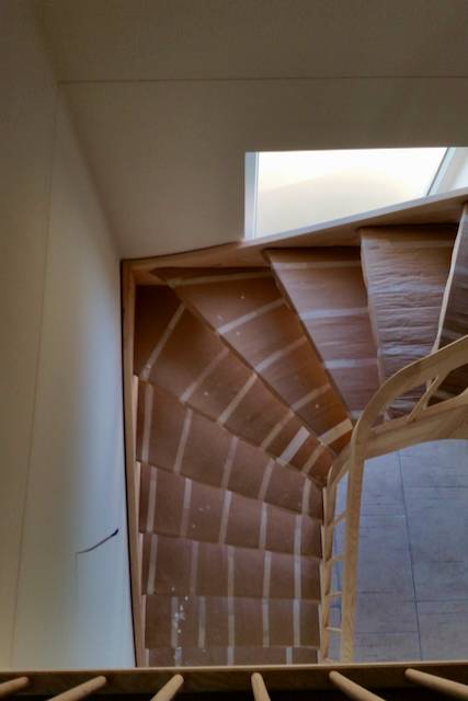 Blick von oben auf eine hölzerne Wendeltreppe mit natürlichem Licht aus einem rechteckigen Oberlicht am oberen Ende, das die geometrischen Linien und Muster der Treppe und des Geländers hervorhebt.
