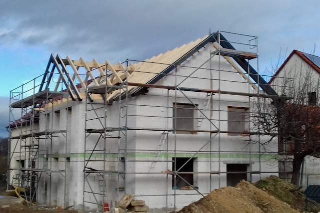 Ein zweistöckiges, im Bau befindliches Haus mit einem Gerüst drum herum und freiliegenden Dachbalken; die Wände sind unvollendet, und Baumaterialien liegen in der Nähe verstreut.