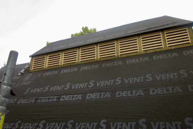 Nahaufnahme eines im Bau befindlichen Gebäudes mit einer Reihe gelber Lüftungslamellen in der Nähe des Daches und schwarzem wetterfestem Material mit der Aufschrift DELTA VENT S an den Außenwänden.